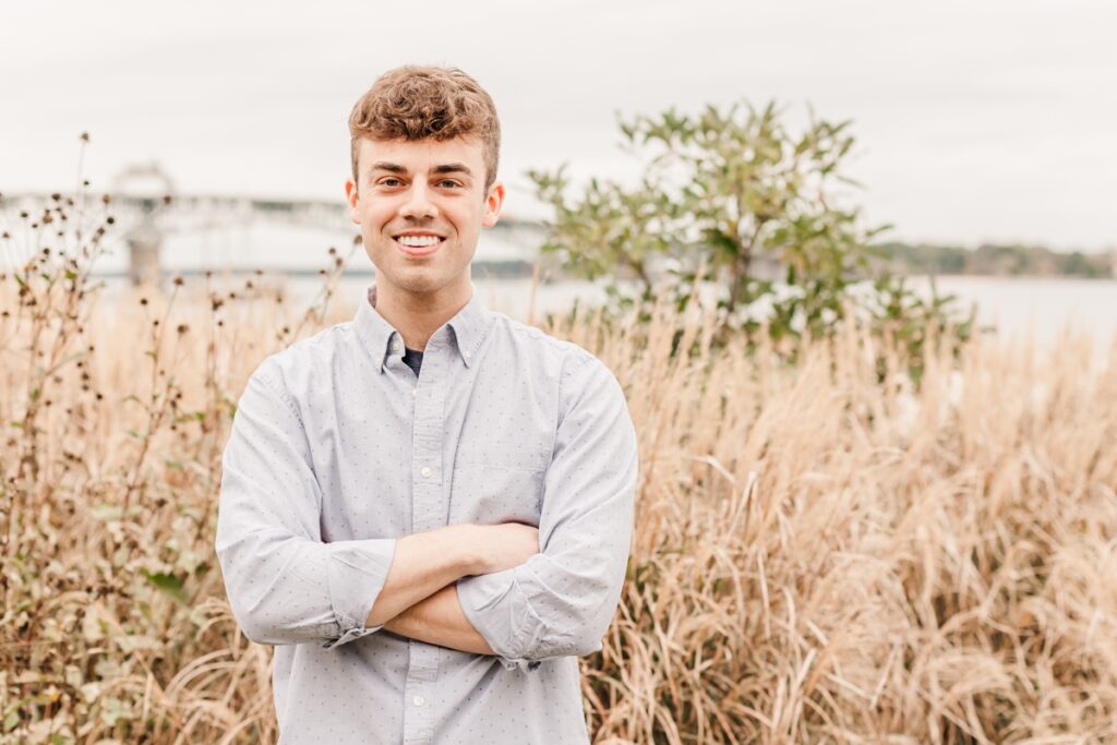 A male graduate smiling at the beach with sea grass blurred in the background.
