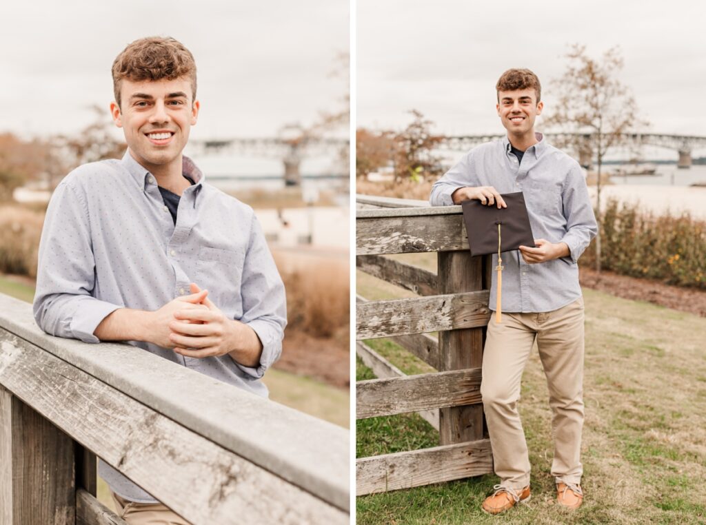 A male graduate in casual attire holding his graduation cap leaning against a fence at the beach. 